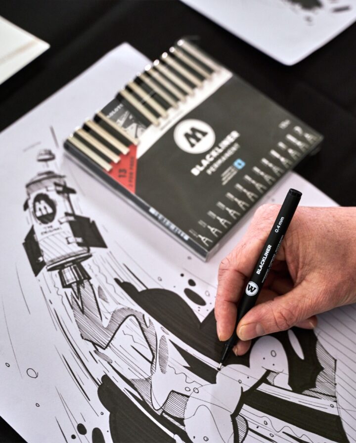 Hand mit schwarzem Fineliner malt ein detailliertes Artwork im Comic-Stil vor einem liegenden Fineliner-Komplettset
