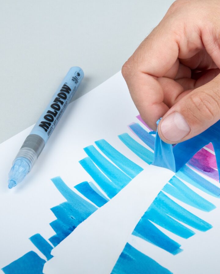 Marker mit Maskierflüssigkeit liegt neben einer Hand, welche die getrocknete Maske vom blau angemalten Papier abzieht