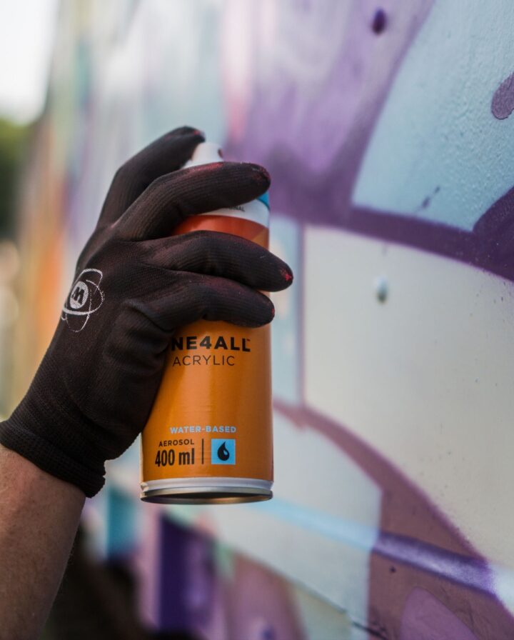 Hand in schwarzem Handschuh sprüht mit orangener Spraydose an eine Wand mit lila Graffiti