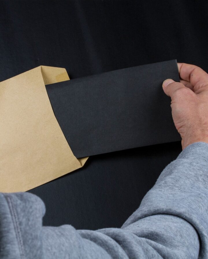 Foto auf dem ein Mann einen befüllten schwarzen Umschlag in einen braunen, großen Umschlag steckt
