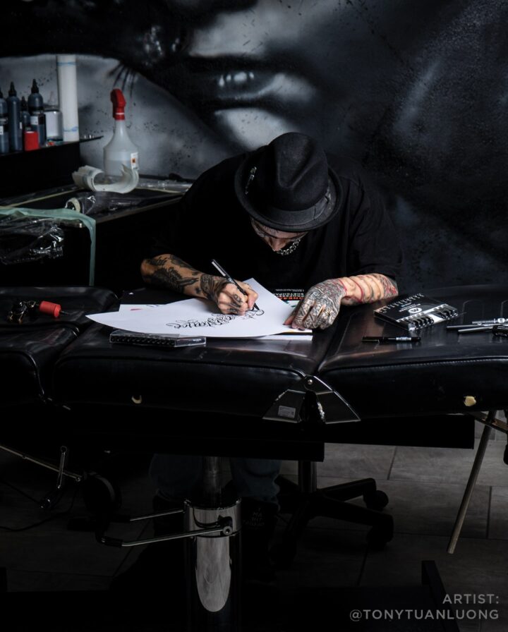 tätowierter Künstler mit schwarzem Hut lehnt auf einer Liege und kalligrafiert einen Schriftzug mit einem dünnen schwarzen Stift
